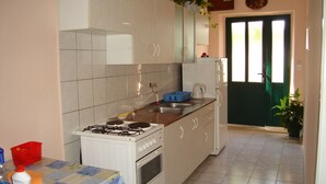 Una cocina funcional con estufa a gas, horno y lavamanos. Se observa una mesa con mantel y una silla. En la esquina, hay una planta en maceta.