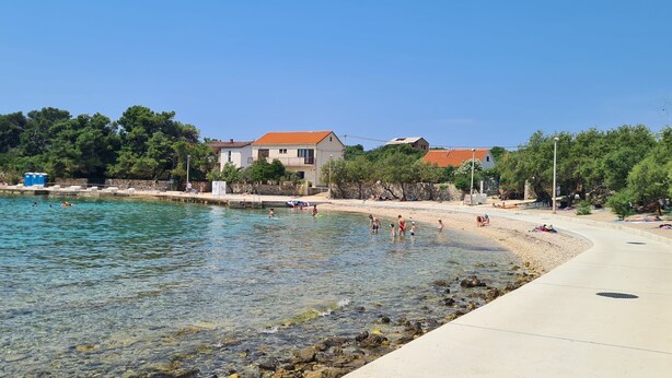 Una zona costera con agua cristalina, una playa arenosa y algunos edificios al fondo.