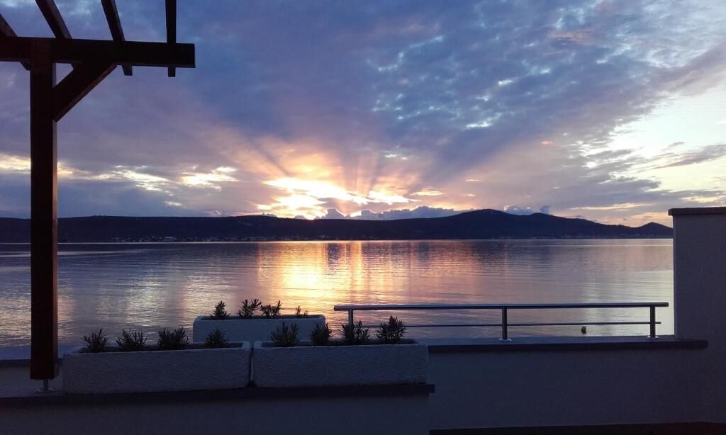 Pôr do sol sobre um lago calmo, com montanhas ao fundo, uma pérgola e um corrimão com plantas.