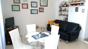 Una sala de estar moderna con una mesa de comedor de vidrio, sillas blancas, un sofá negro y un televisor sobre un mueble de madera.