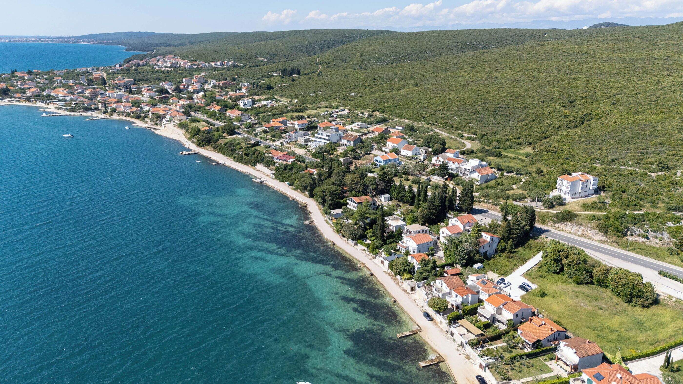 An aerial view of a coastal town with houses along the beach, clear blue water, and green hills in the background.