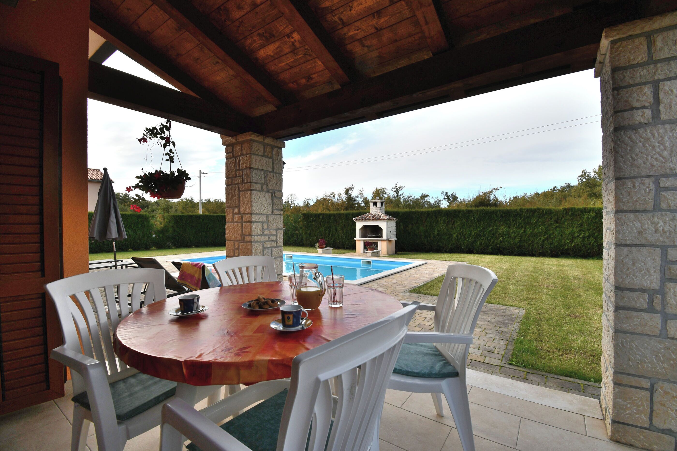 Un porche cubierto con una mesa de madera y sillas blancas, con vistas a una piscina y jardín.
