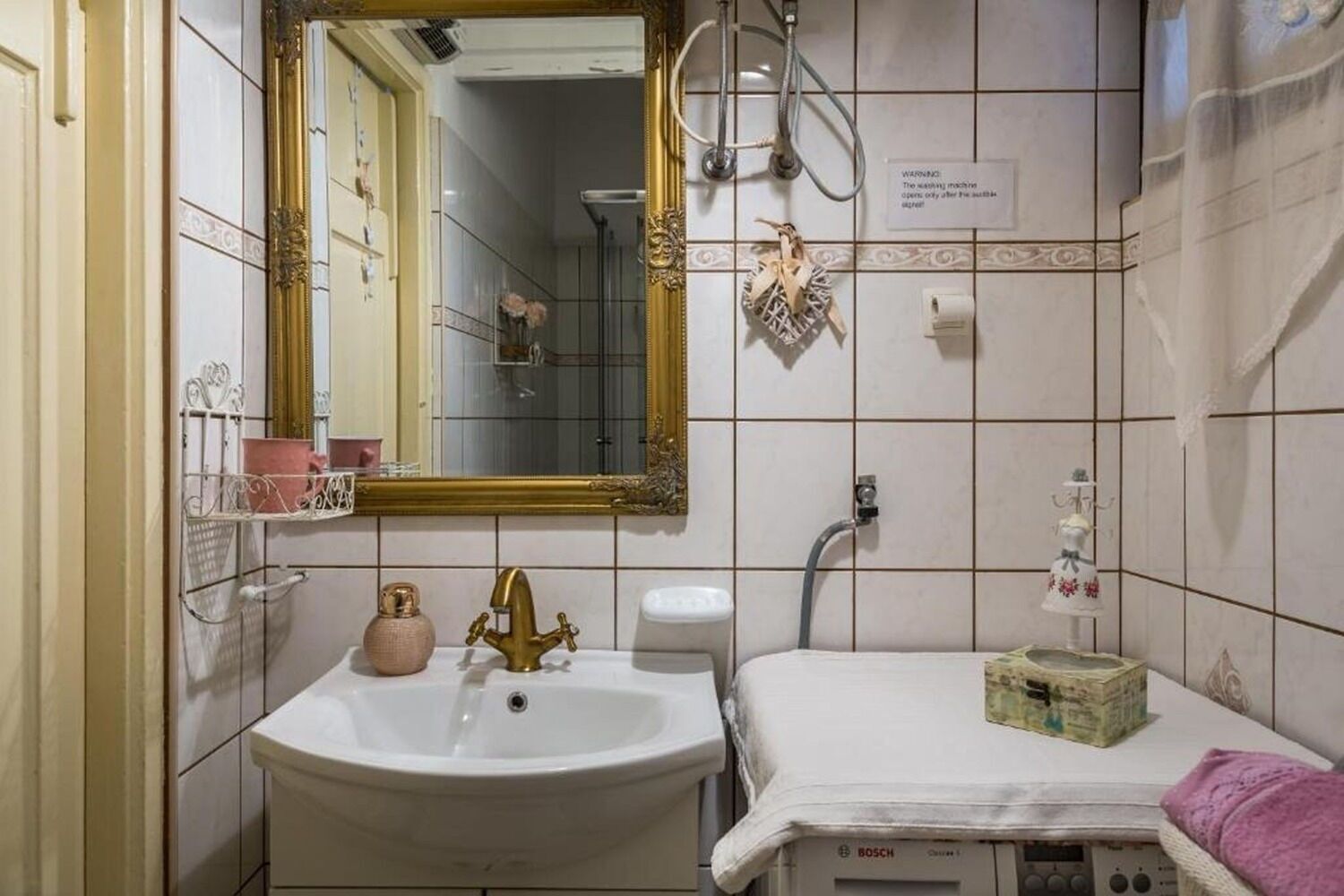Een badkamer met een witte wastafel, een gouden kraan en een spiegel met een gouden lijst. Er is een douchegedeelte met een douchekop en een handdoekhouder.