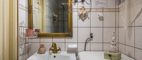 Een badkamer met een witte wastafel, een gouden kraan en een spiegel met een gouden lijst. Er is een douchegedeelte met een douchekop en een handdoekhouder.