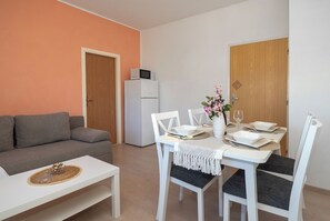 Ein modernes Wohnzimmer mit einem weißen Sofa, einem Esstisch mit weißen Stühlen, einem weißen Kühlschrank und einer Holztür.
