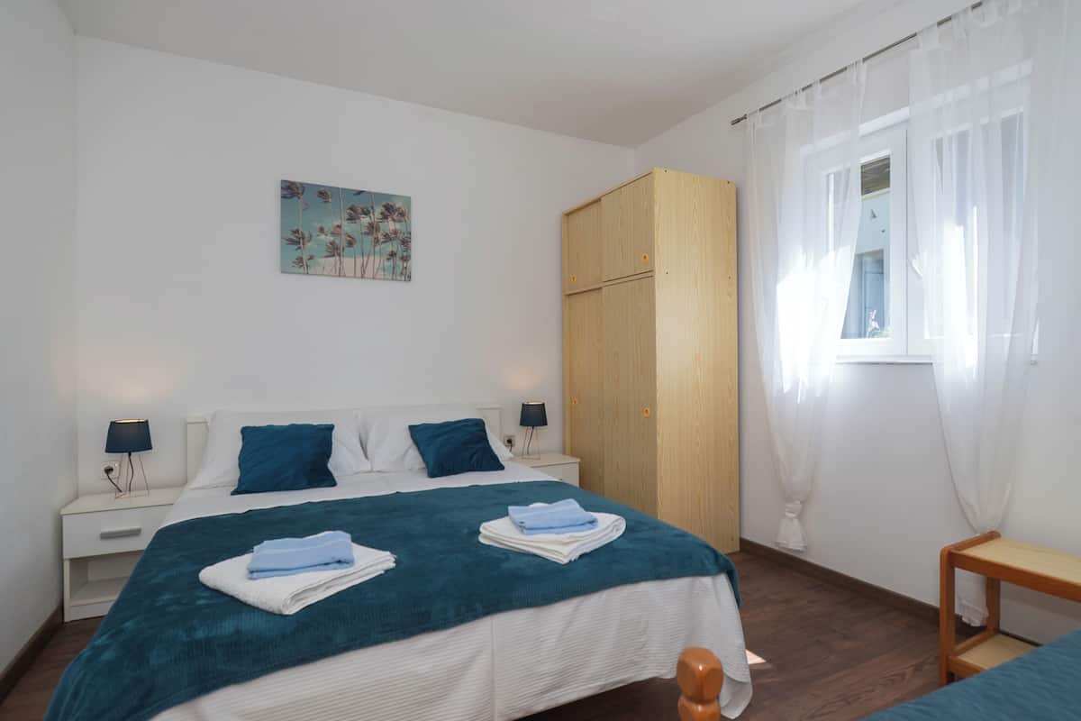 Un dormitorio con cama, mesitas de noche, un armario de madera, una ventana con cortinas y un cuadro en la pared.