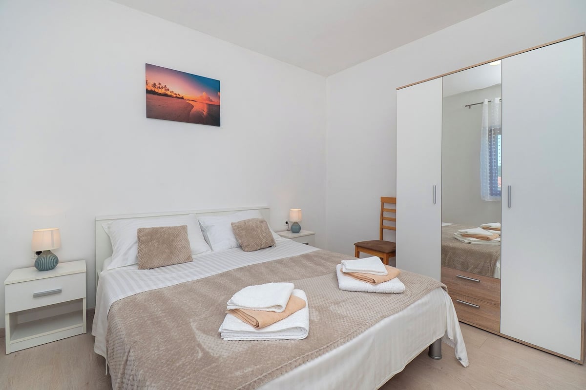 Un dormitorio moderno con una cama, mesitas de noche, una silla, un espejo grande y una pintura de atardecer.