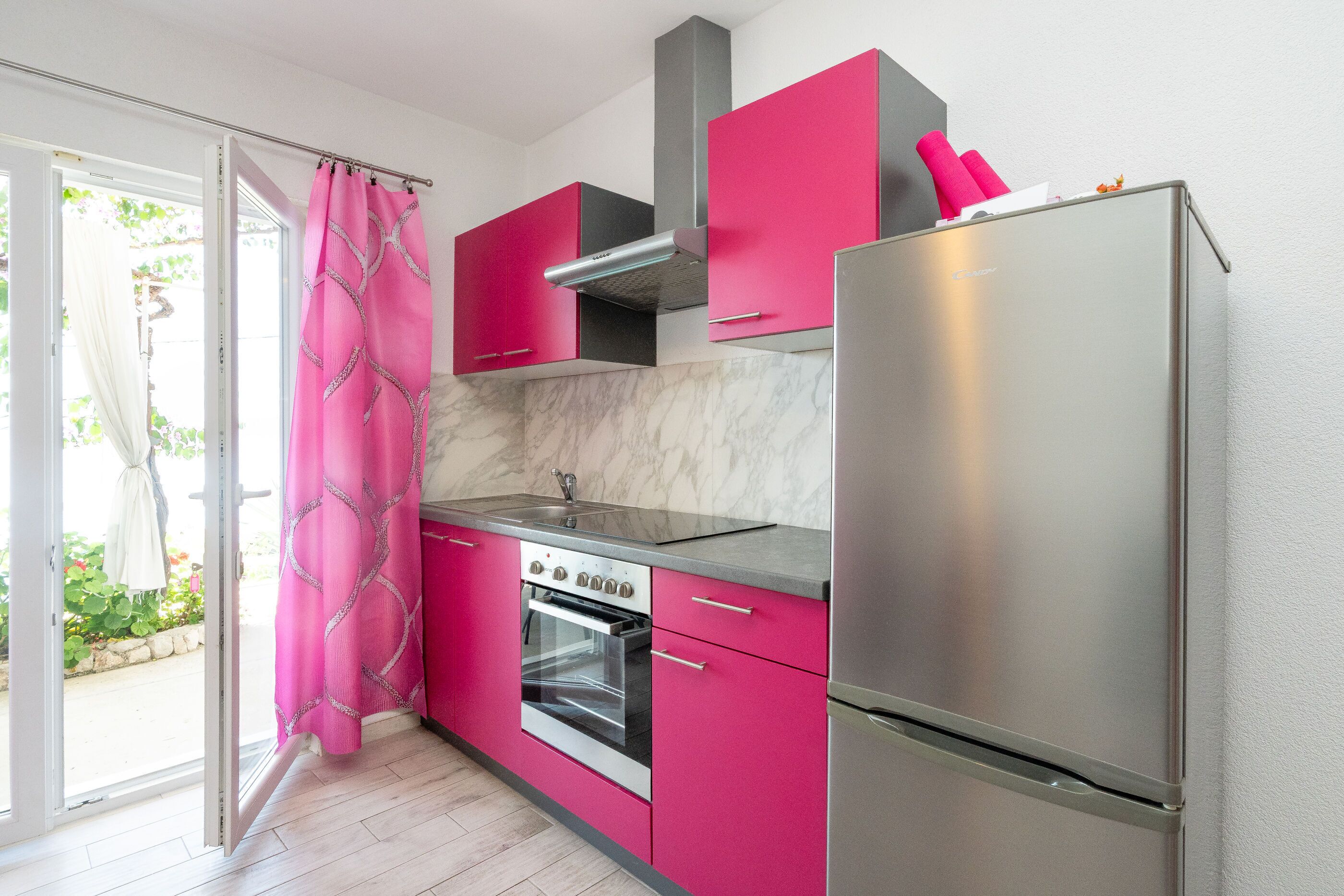 A kitchen with pink cabinets, a stainless steel refrigerator, and a window with a view of greenery.