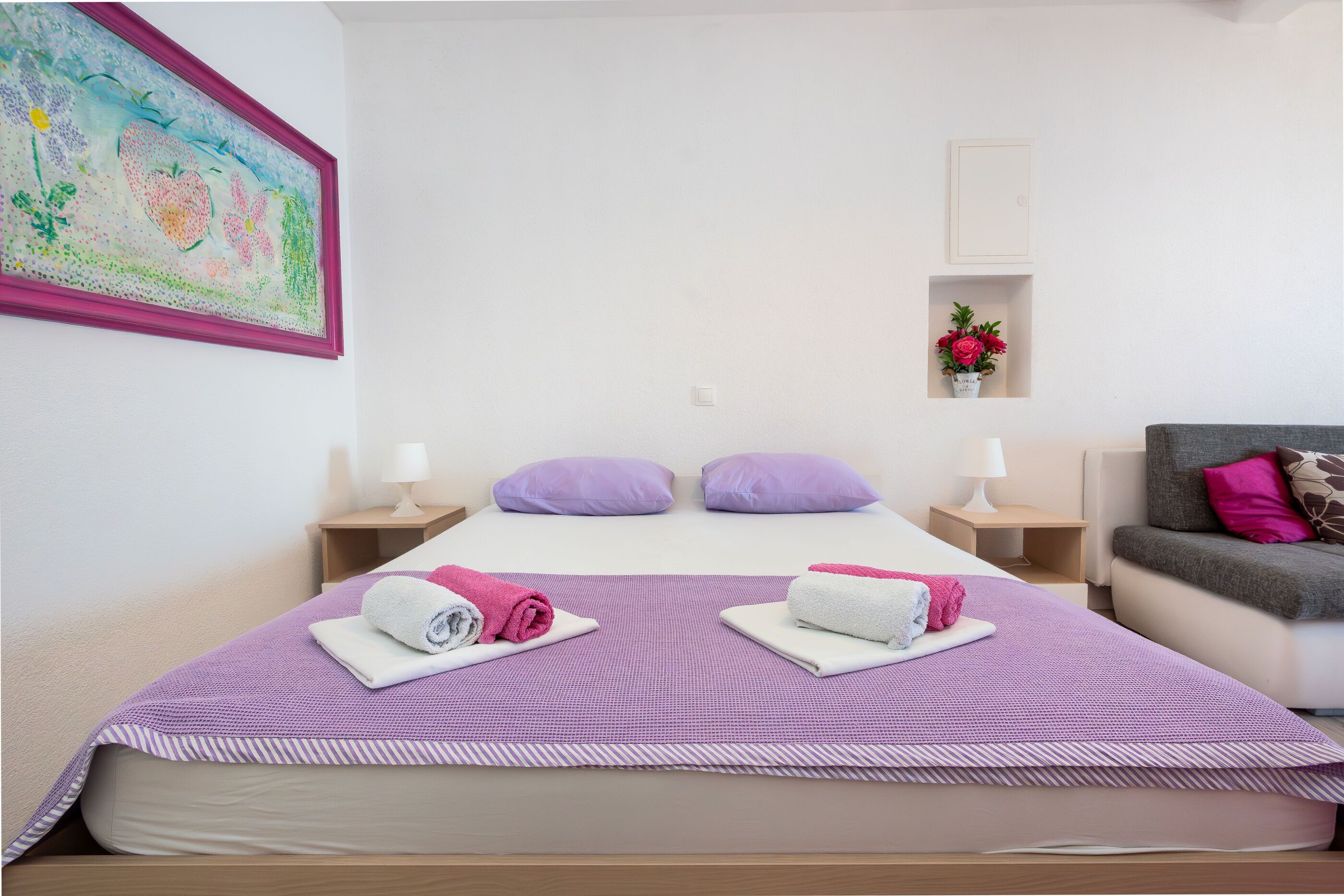 Habitación de hotel con una cama, dos almohadas, una mesita de noche con una lámpara, un sofá y un estante en la pared con flores.