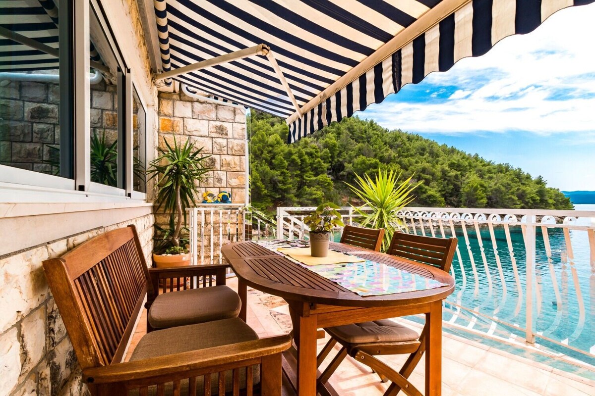 Ein Holztisch mit Stühlen steht auf einem Balkon mit einem Streifenverdeck, mit Blick auf einen Pool und einen bewaldeten Hügel.