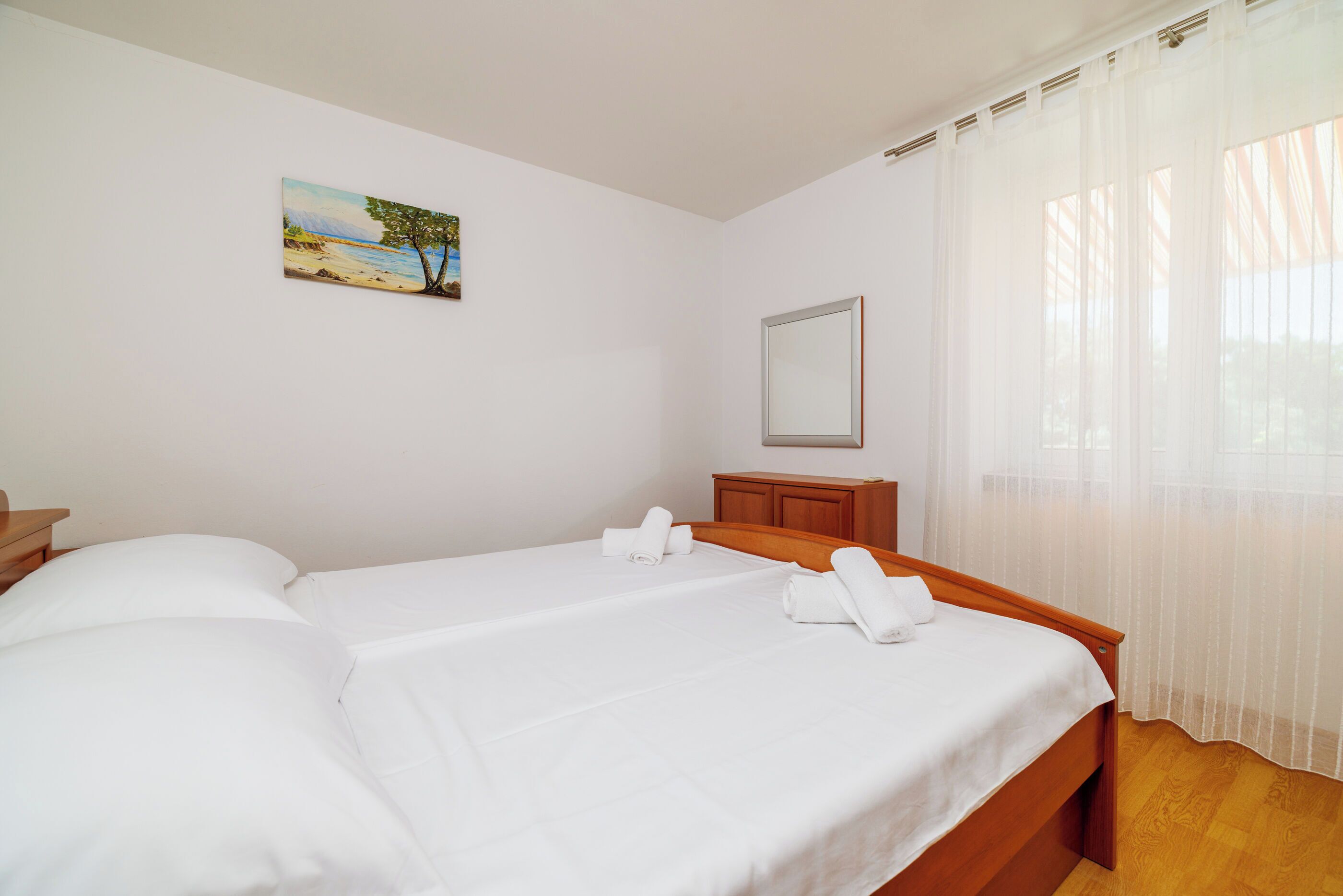 A hotel room with two beds, a wooden headboard, a painting on the wall, and a window with sheer curtains.