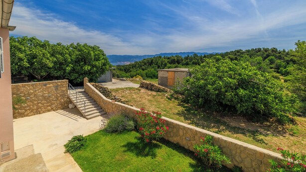 Un espace extérieur délimité par des murs de pierre, avec un escalier menant à un bâtiment, le tout entouré d’une végétation luxuriante et sous un ciel dégagé.