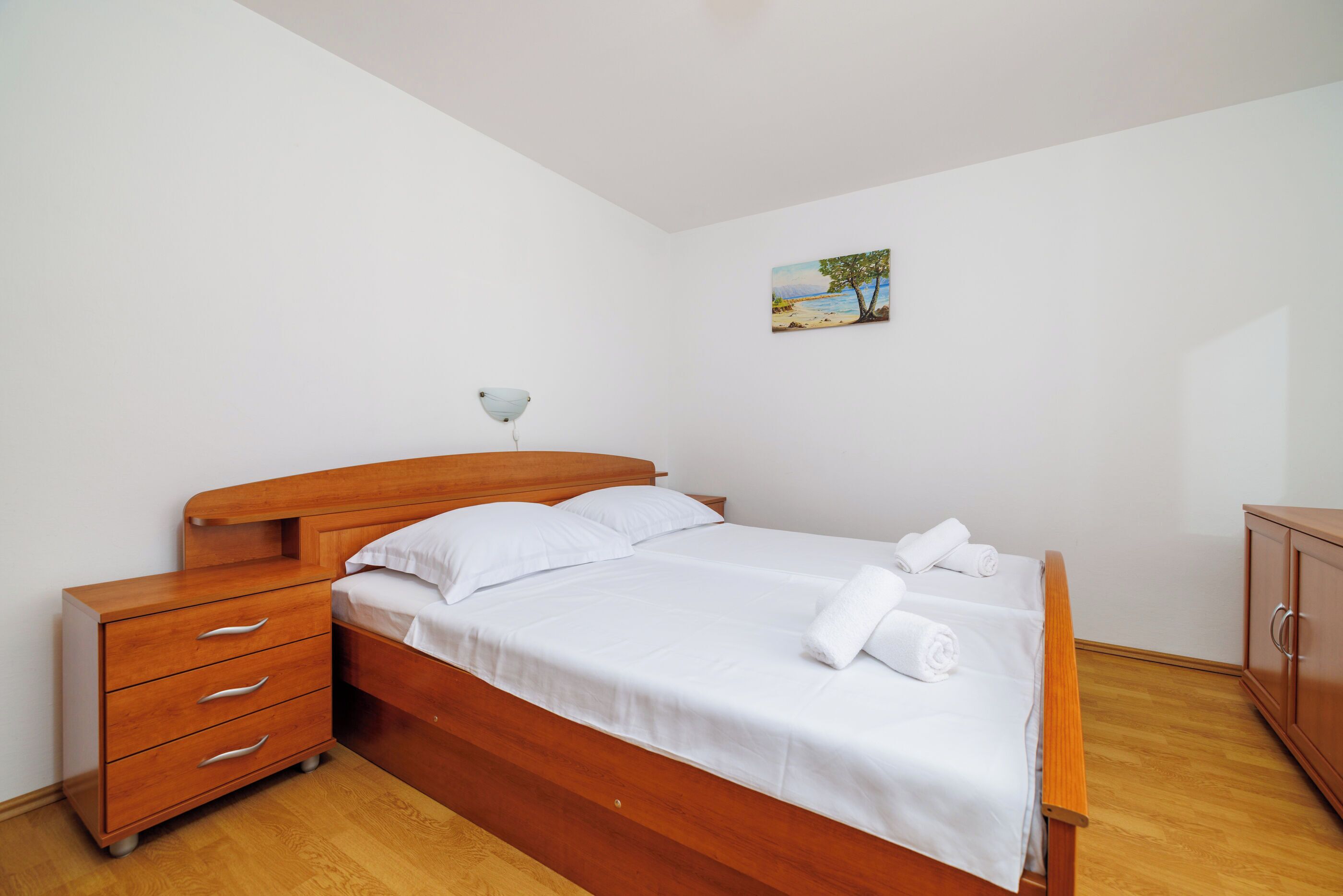 Ein ordentlich bezogenes Bett mit weißen Leinen und Handtüchern, ein Nachttisch aus Holz mit Schubladen, ein kleiner Beistelltisch mit Lampe und ein Bild an der Wand.