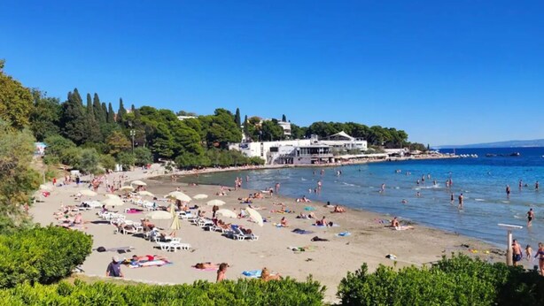 A beach with sun loungers and umbrellas, a building with a white facade, and a clear blue sky. - Apartment Silva (Split)