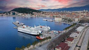 A coastal cityscape with a large ferry docked at a harbor, surrounded by urban buildings and a mountainous backdrop. - Apartment Silva (Split)