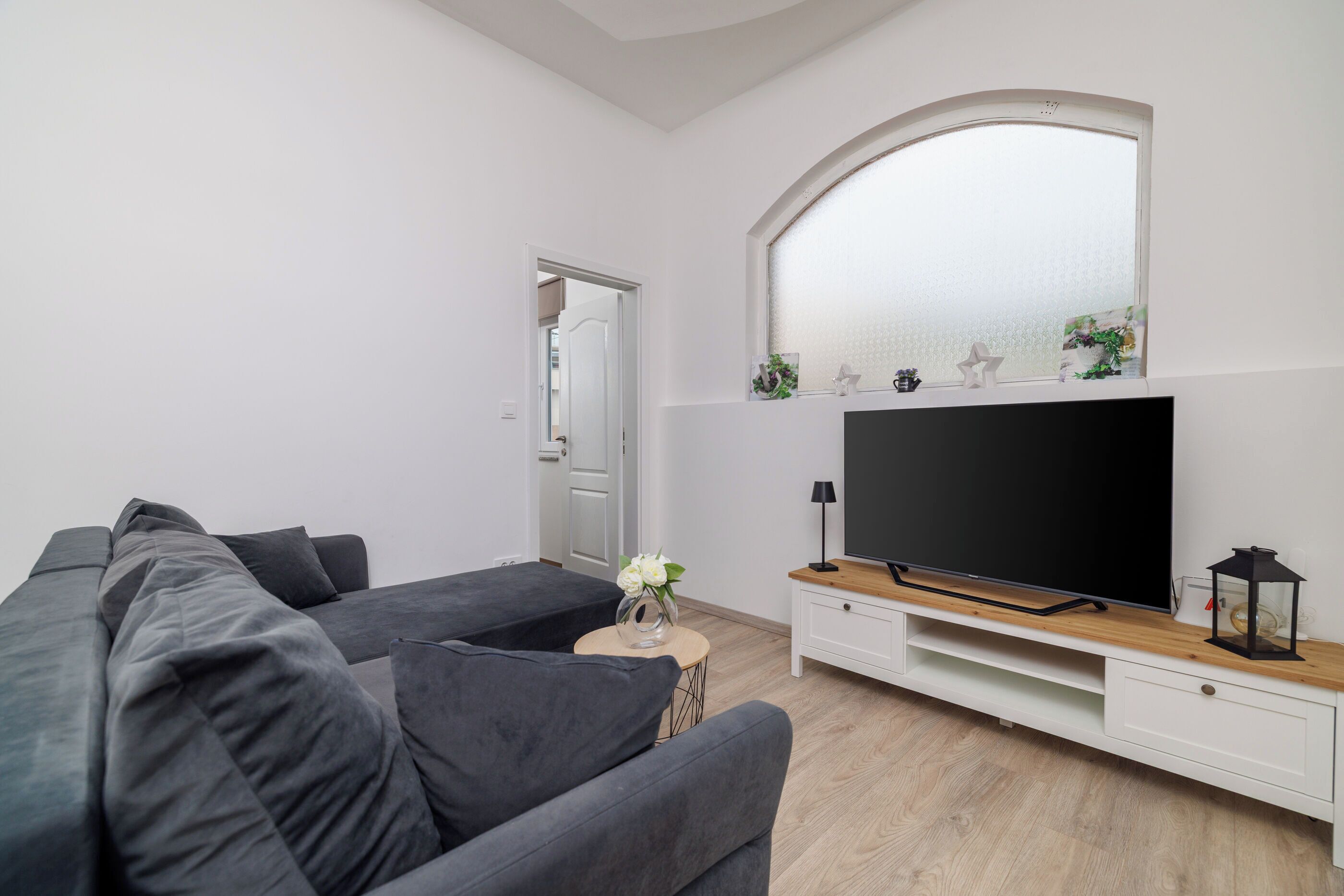 Uma sala de estar moderna com um sofá cinzento, uma televisão de tela plana sobre um suporte de madeira, uma mesinha de canto com um vaso de flores, e uma porta branca ao fundo.