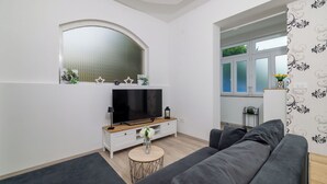 A modern living room with a grey sofa, a TV on a wooden stand, a small round table with a vase, and a large arched window. - Apartment Filip 2 (Crikvenica)