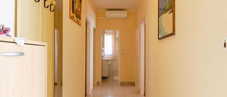 A narrow hallway with a wooden cabinet on the left, a painting on the right, and a door at the end leading to a bathroom.