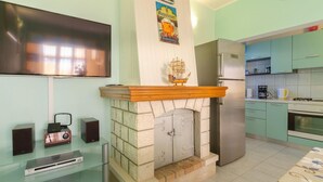 Una sala de estar con chimenea, un televisor de pantalla plana montado en la pared y una cocina con armarios y electrodomésticos de colores claros.