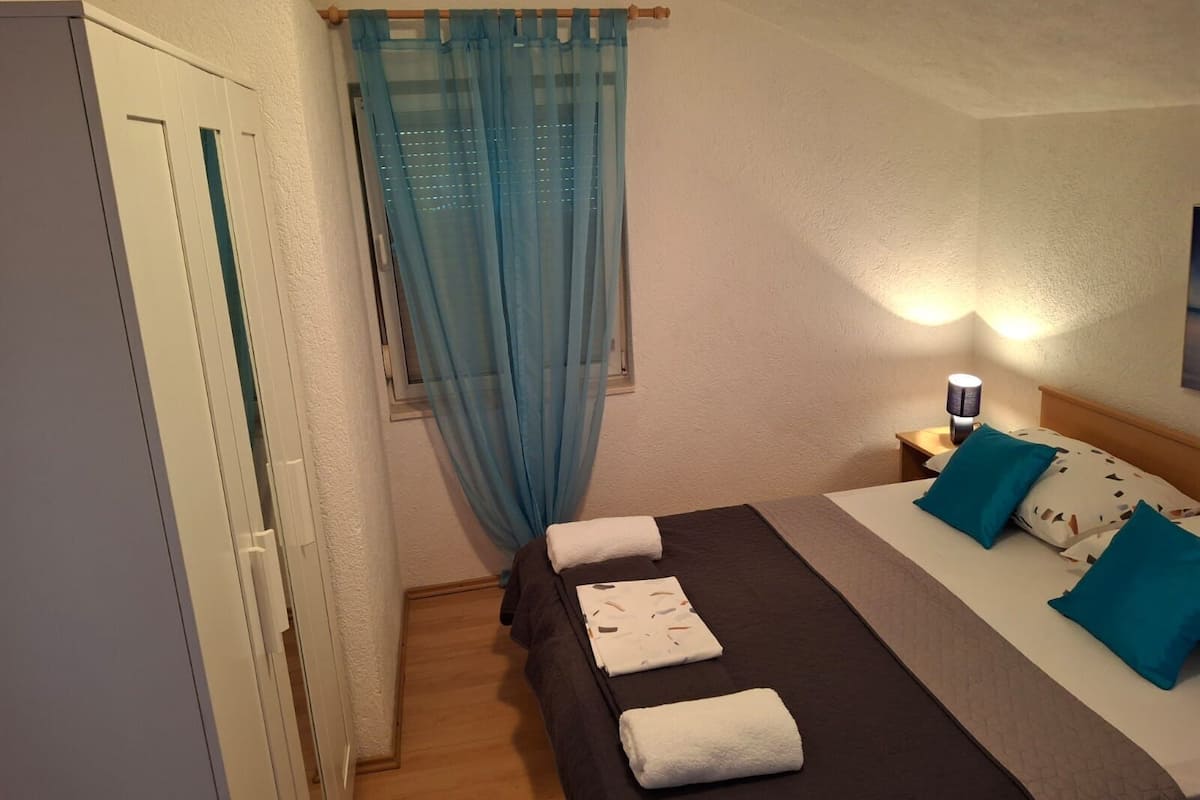 Ein Schlafzimmer mit Bett, Nachttisch, Lampe, Fenster mit leichtem Vorhang und Tür.