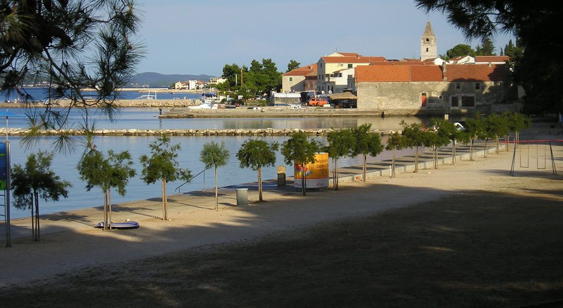 A coastal area with a sandy beach, a row of palm trees, and a building with a red roof in the background.
