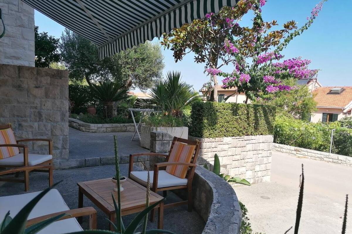 An outdoor seating area with wooden chairs and tables, surrounded by greenery and a stone wall.