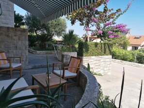 An outdoor seating area with wooden chairs and tables, surrounded by greenery and a stone wall. - Apartments Family (Postira)