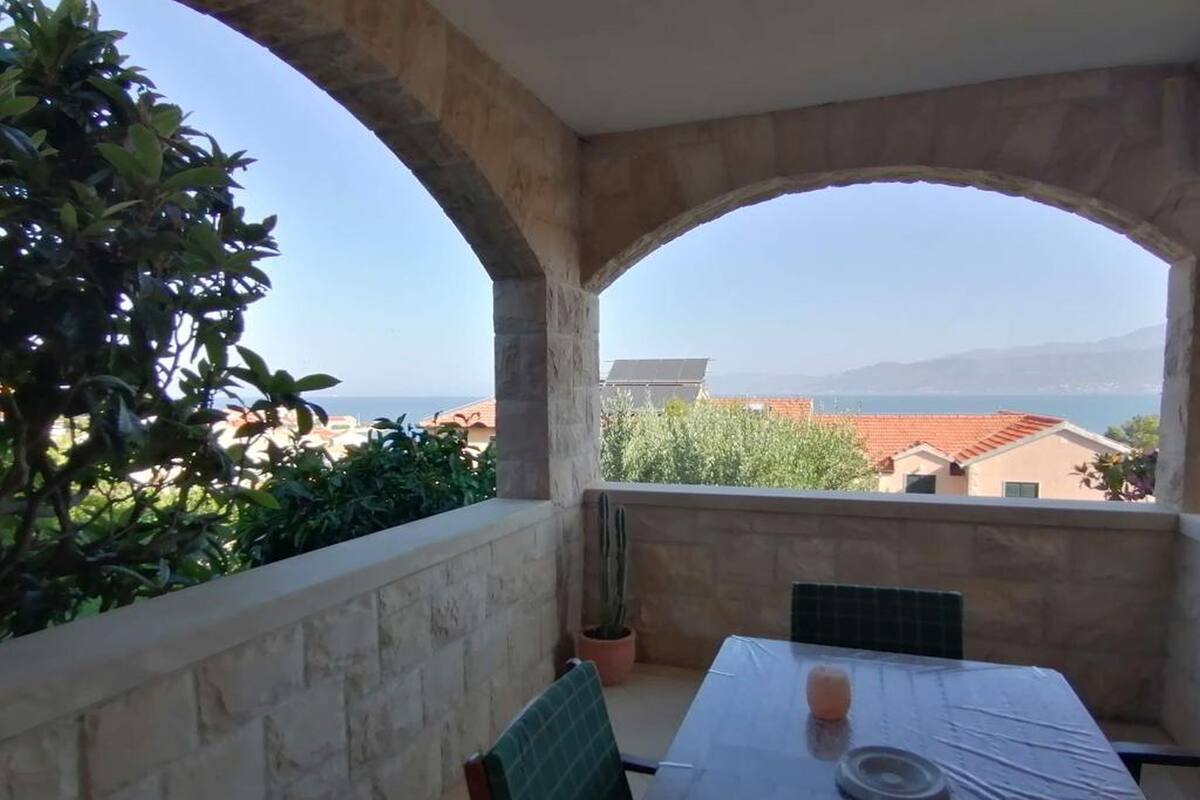 A balcony with a table and chairs, a potted plant, and a view of the sea and buildings.