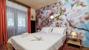 Hotelkamer met een groot bed, een nachtkastje met lamp, bloemen behang en een raam met gordijnen.