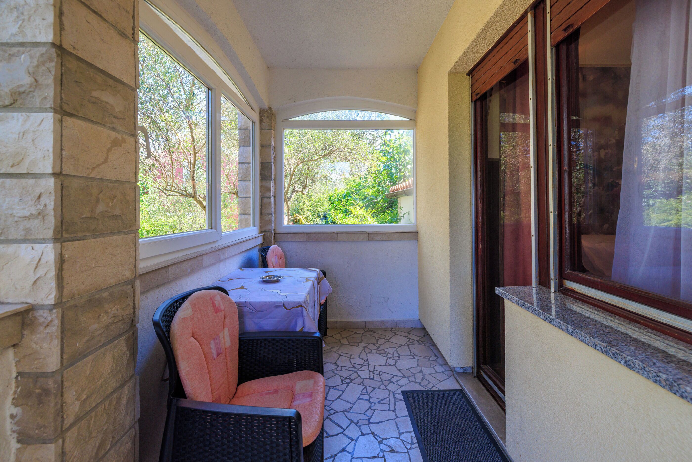 A small balcony with a table and two chairs, a window with a view of greenery, and a door with a curtain.