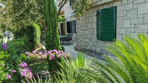 A stone building with green shutters, surrounded by lush greenery and flowering plants. - Apartment Vesna (Postira)