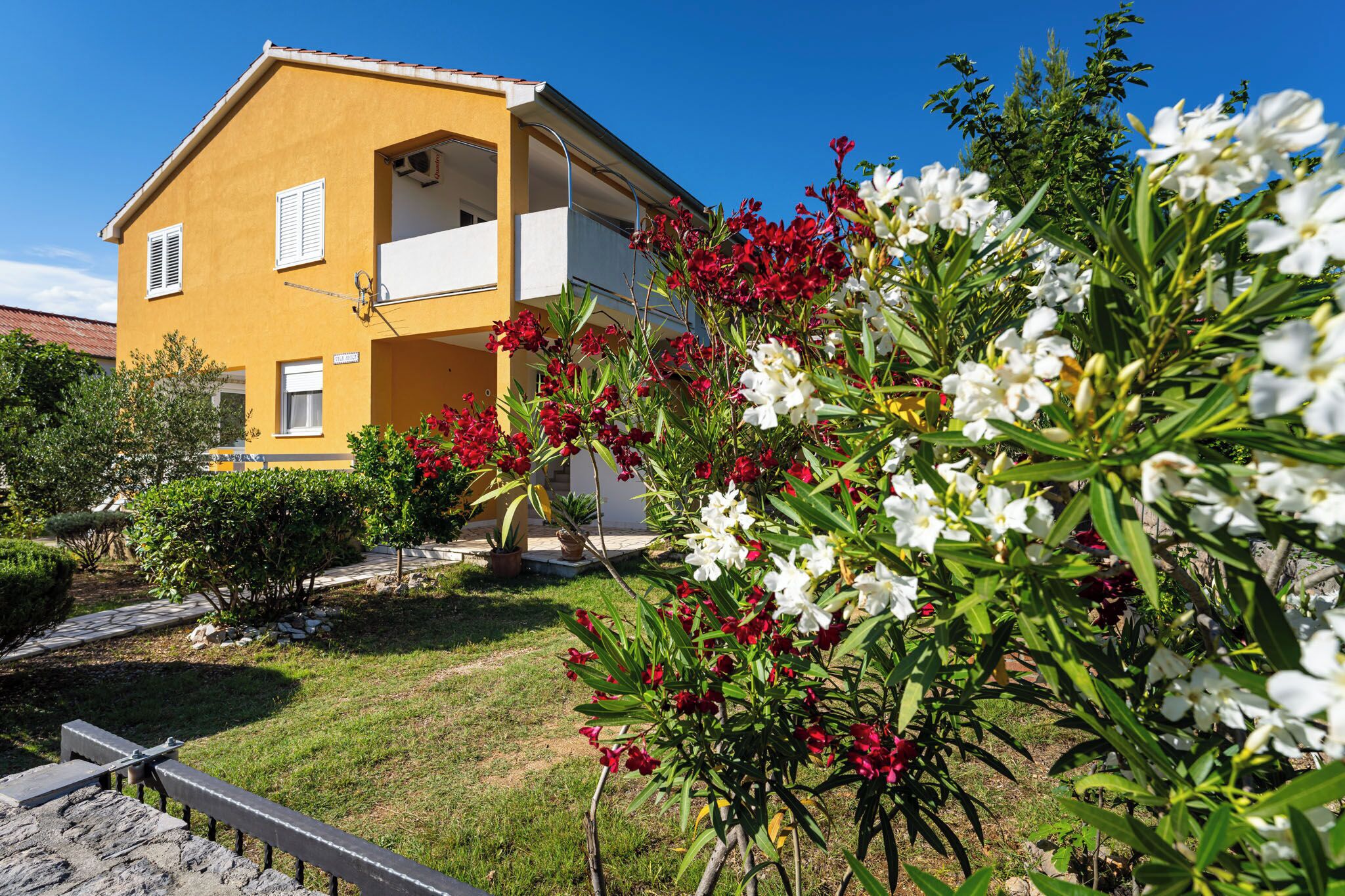Un bâtiment jaune à deux étages, doté d’un balcon, entouré d’un jardin où poussent des fleurs rouges et blanches.