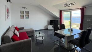 A modern living room with a sofa, a glass coffee table, a dining table with chairs, a TV, and a view of the outdoors. - Apartments Dobrila (Jelsa)