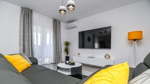 Egy modern nappali, szürke kanapéval, sárga párnákkal, kerek dohányasztallal, televízióval és állólámpával.