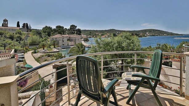 A balcony with two green chairs overlooking a coastal area with a view of the sea, a church, and a stone building. - Apartment Milan (Trogir)