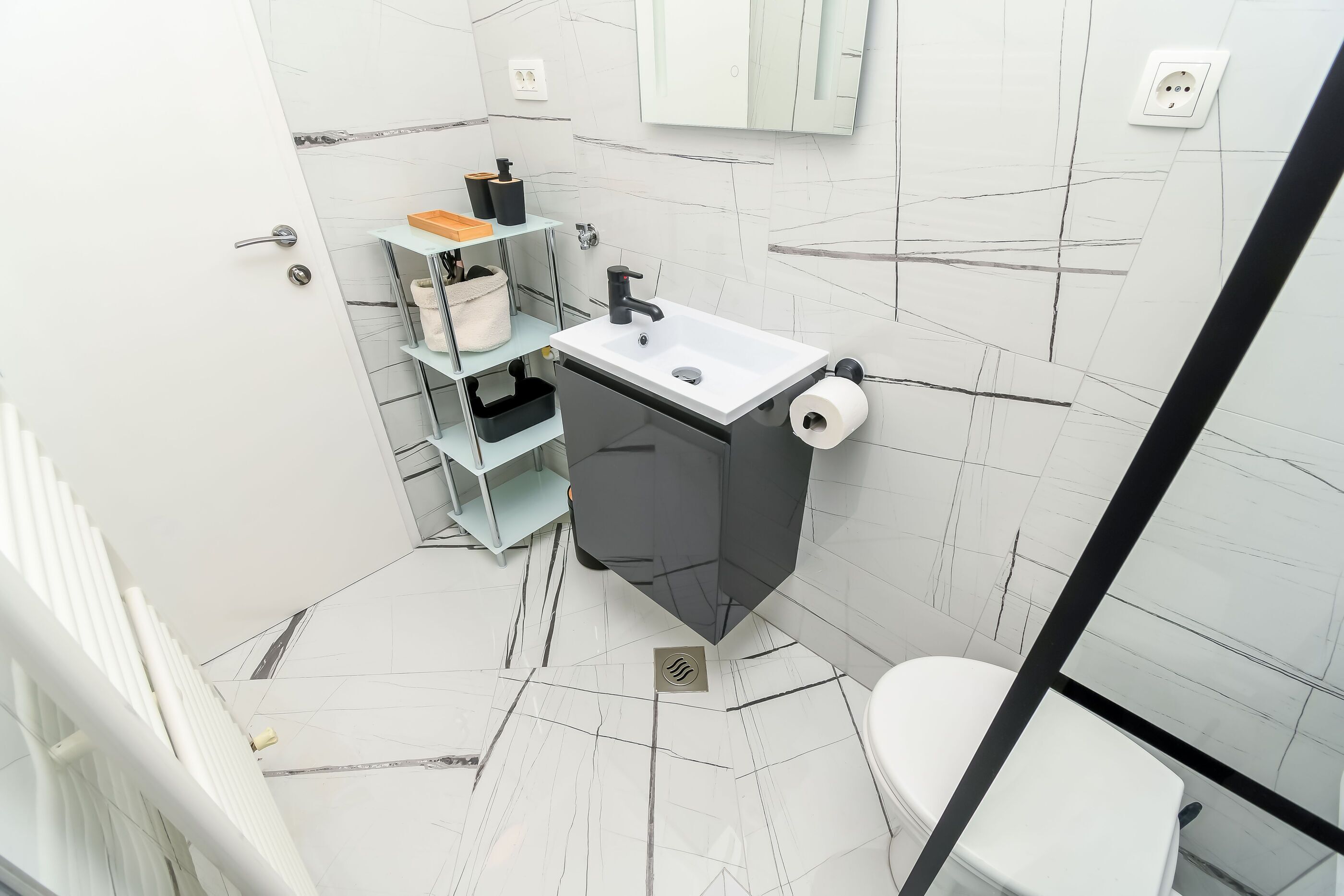 Un baño pequeño con un lavamanos blanco, un mueble negro y un inodoro. Hay repisas con artículos de aseo y un espejo sobre el lavamanos.