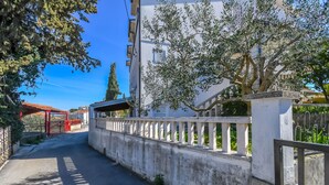 Ένας στενός δρόμος με μια λευκή περίφραξη και μια κόκκινη πύλη, που οδηγεί σε ένα κτίριο με λευκή πρόσοψη και ένα δέντρο μπροστά.