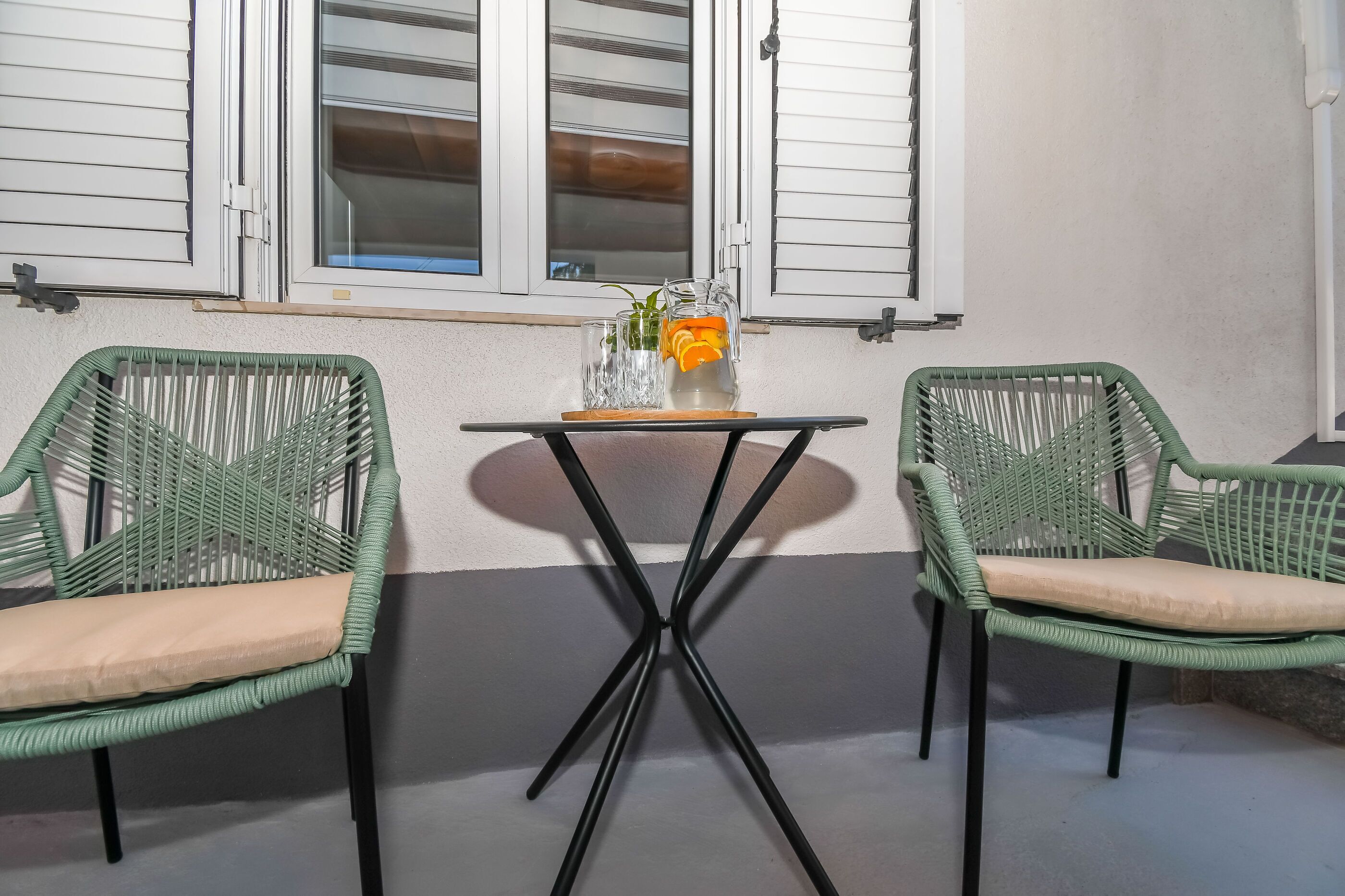 Dos sillas verdes de mimbre con cojines beige, una mesita con un tarro de cristal y rodajas de naranja, y una ventana con contraventanas blancas.