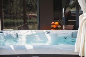 A hot tub with a view of a forest, a wooden table with fruit, and a chair.