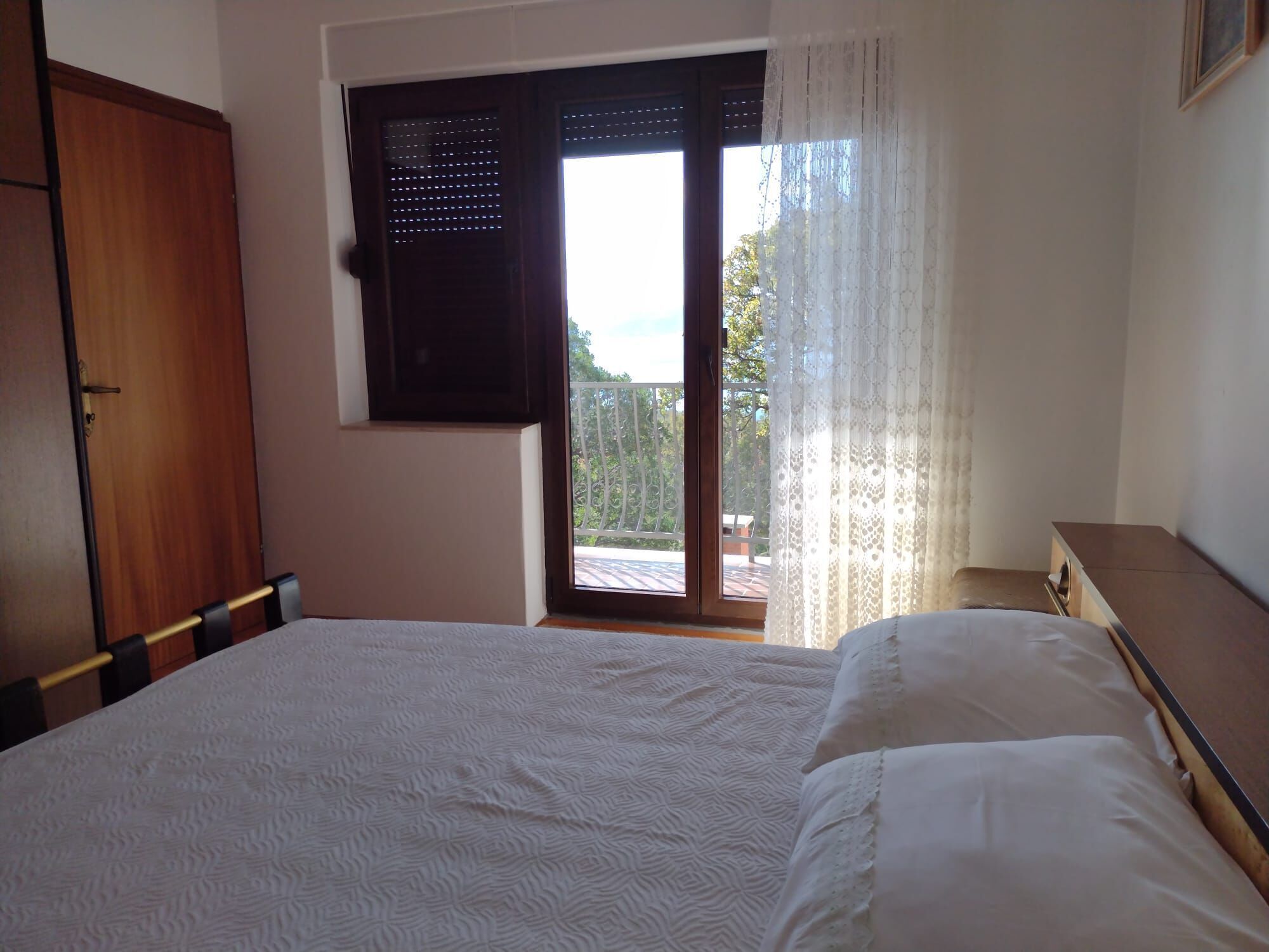 A bedroom with a bed, a wooden wardrobe, a window with a view of the outdoors, and a balcony.