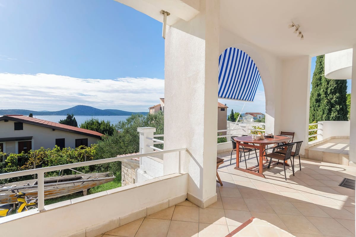 A balcony with a table and chairs, overlooking a body of water and distant mountains.