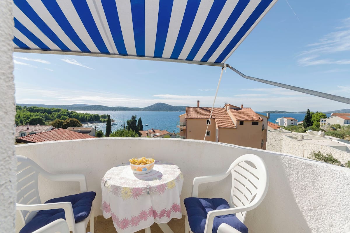 A balcony with a table and chairs, overlooking a coastal view with houses and a body of water.