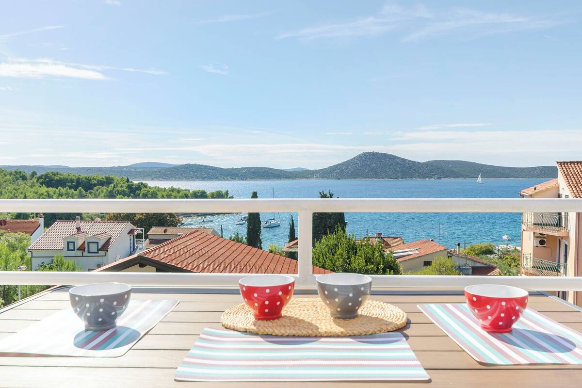 A balcony with a view of a lake and mountains, featuring two tables with striped mats and polka-dotted bowls.