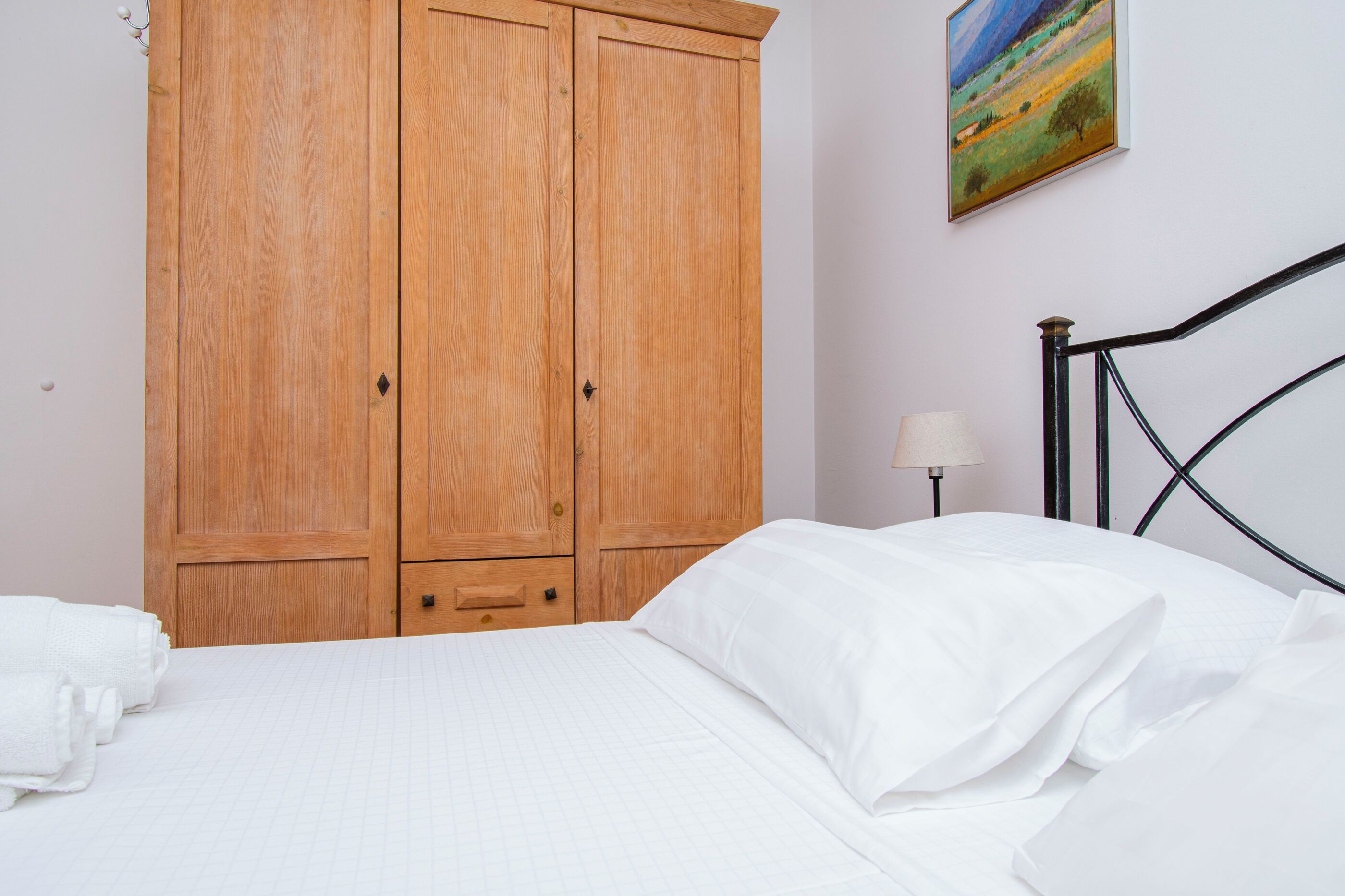 A bedroom with a bed, a wooden wardrobe, a painting on the wall, and a nightstand with a lamp.