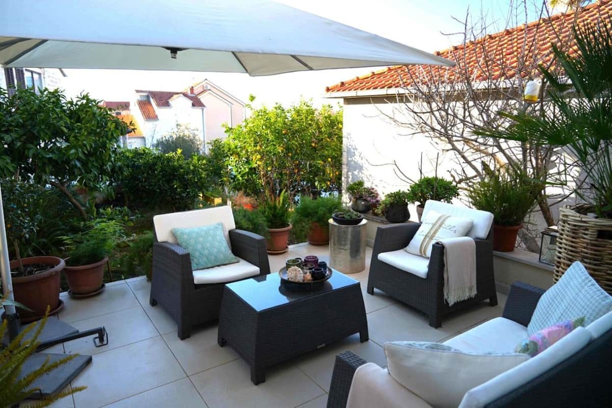 An outdoor patio area with wicker furniture, potted plants, and a large umbrella providing shade.