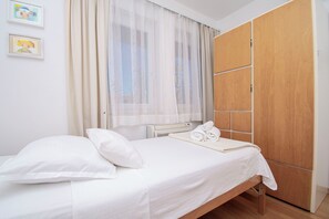 A bedroom with a bed, a window with sheer curtains, a radiator, and a wooden wardrobe. - Apartments Apartmani Oh La La (Supetar)
