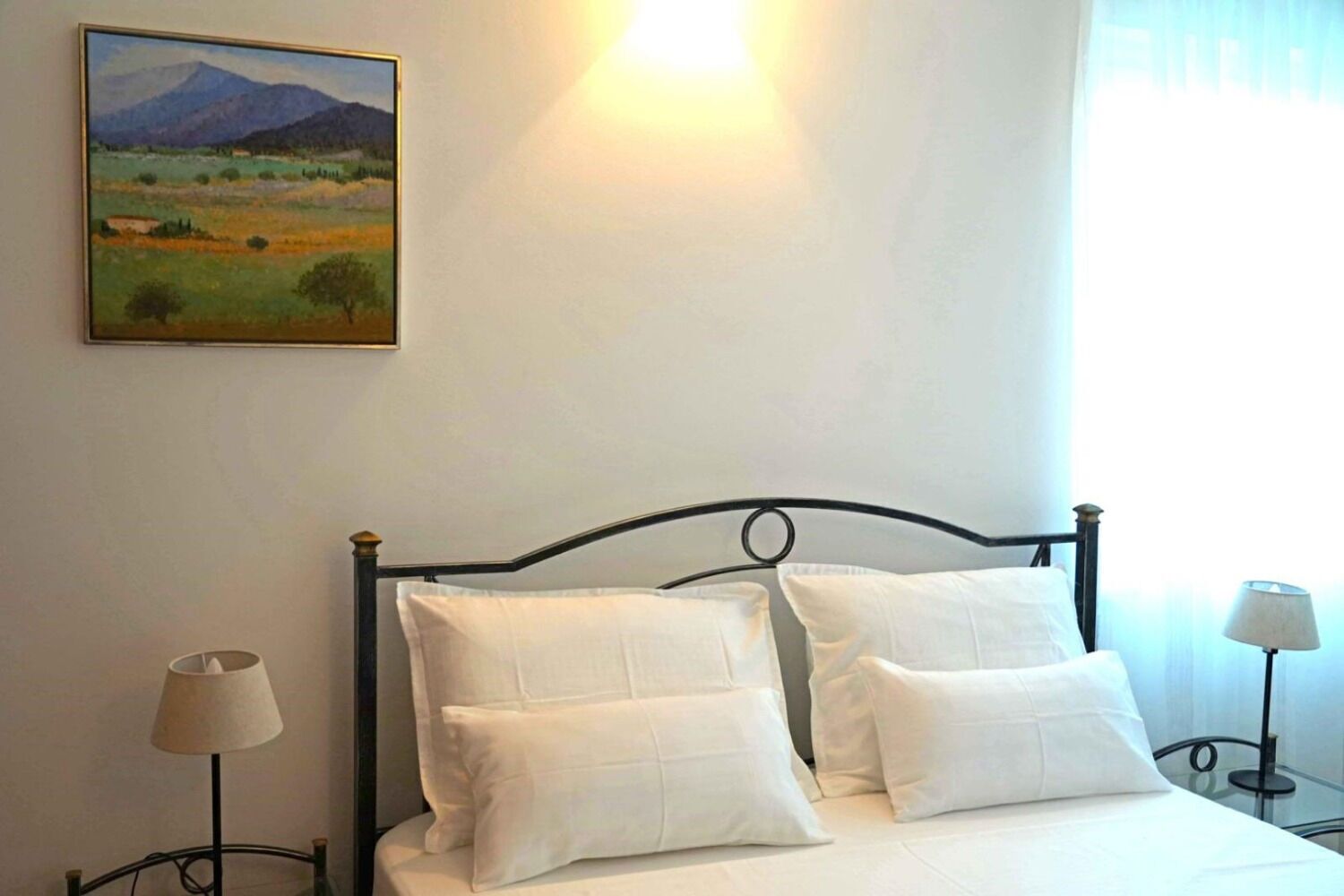 A bed with white bedding and two pillows, a painting of a landscape on the wall, and two bedside lamps.
