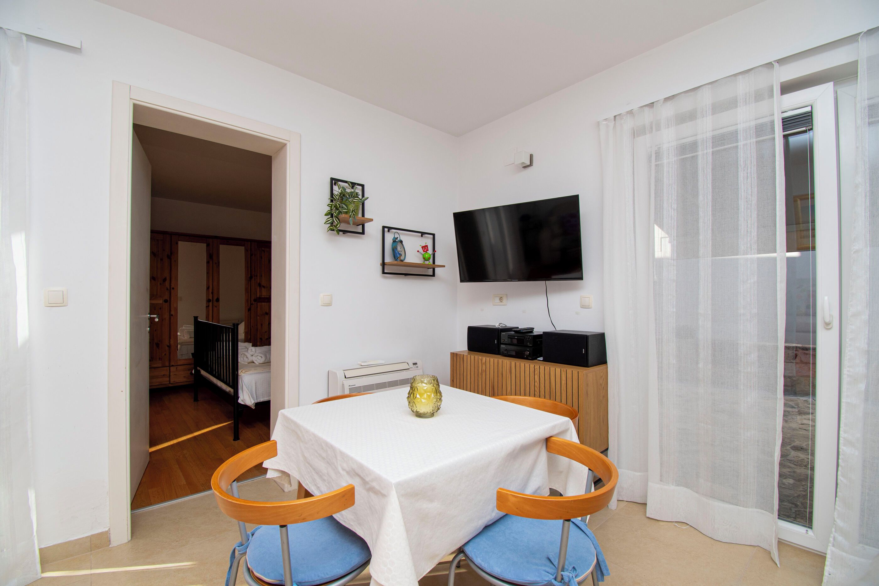 A small, well-lit room with a dining table, two chairs, a flat-screen TV, and a window with sheer curtains.