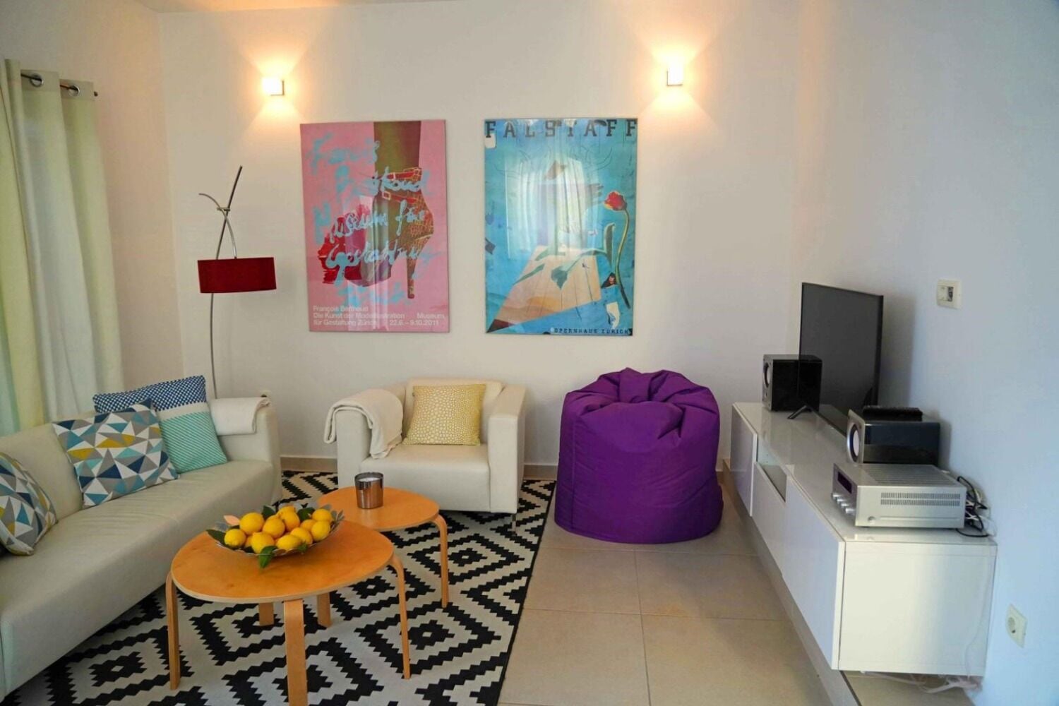 A living room with a sofa, coffee table, bean bag, and a TV. There are two posters on the wall and a lamp.