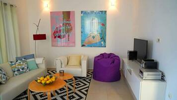 A living room with a sofa, coffee table, bean bag, and a TV. There are two posters on the wall and a lamp.